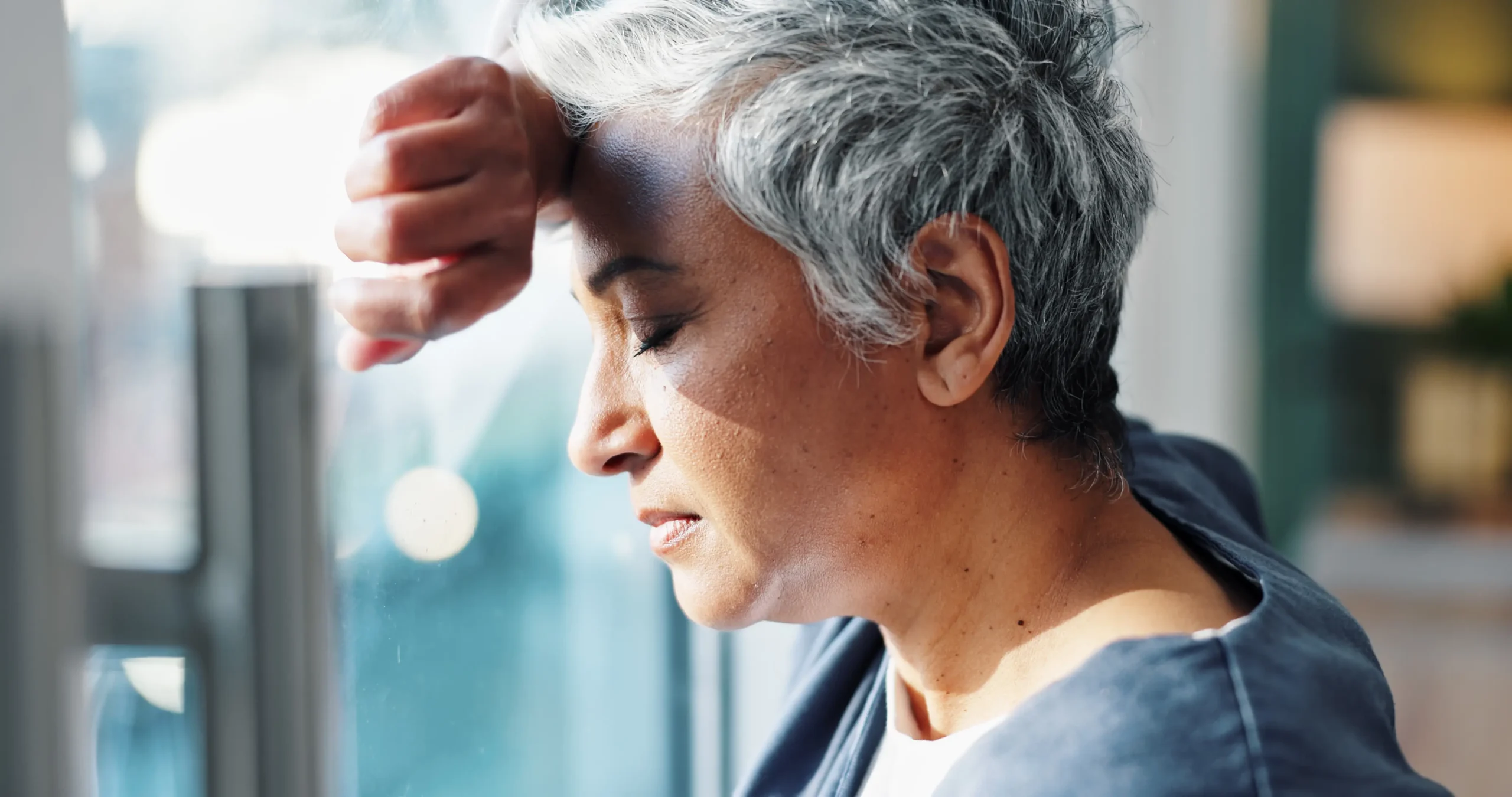 Mature woman leaning her head against a hospital window, appearing exhausted and burned out, representing caregiver fatigue and mental health challenges.